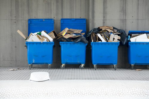 A row of five large blue wheeled rubbish bins positioned on a paved pavement beside a grey concrete wall, with each bin filled with various types of discarded waste including cardboard boxes, paper, and plastic items. The contents appear to be somewhat overflowing, with some waste spilling over the top edges, and one white piece of paper lying on the ground in front of the bins. The environment suggests an outdoor urban or commercial area in Chiswick, where waste collection and rubbish removal services by Chiswick House Clearance might operate, highlighting typical waste disposal methods for household or business waste in the locality.