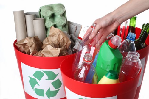 A close-up image of two red recycling bins, each marked with a green recycling symbol, filled with various recyclable waste materials. The left bin contains several brown crumpled paper bags, multiple cardboard tubes, and a few empty plastic egg cartons made of greenish foam. The right bin holds an assortment of plastic bottles, containers, and jars, including clear, green, and red-tinted plastic bottles, some with caps on, along with a yellow plastic bottle. A person's hand wearing a transparent glove is seen placing or retrieving a plastic bottle from the right bin. The setting appears to be outdoors, possibly on a driveway or paved area, with natural daylight illuminating the scene. The overall context suggests a professional rubbish or recycling collection service, such as Chiswick House Clearance, operating in the local area, aiming to promote proper waste segregation and recycling practices.
