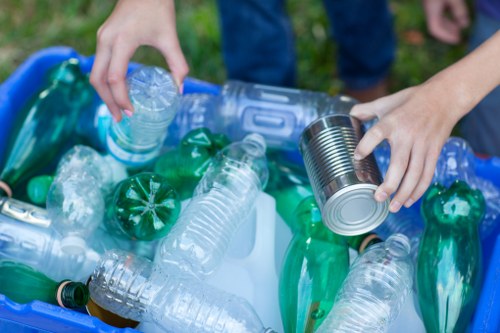 A person's hand is seen placing a plastic bottle into a blue recycling bin filled with various waste plastic bottles, including clear, green, and blue containers, some with caps on. The scene is set outdoors on a grassy area, with part of the person's blue jeans visible. The waste items are piled in the bin, which appears to be part of a rubbish removal or recycling collection, consistent with services offered by Chiswick House Clearance. The environment suggests an active waste disposal or recycling process, with focused attention on properly segregating plastic waste for disposal or recycling in Chiswick or nearby areas.